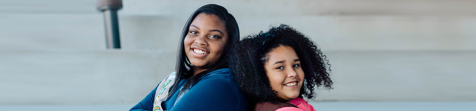  a high school ambasador girl scout standing back-to-back with a young brownie girl scout in elementary school 