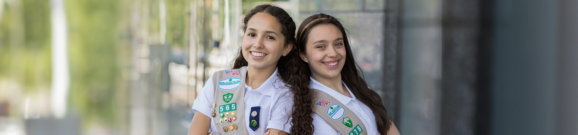  two senior girl scouts wearing sash uniforms outside standing back to back and smiling at the camera 